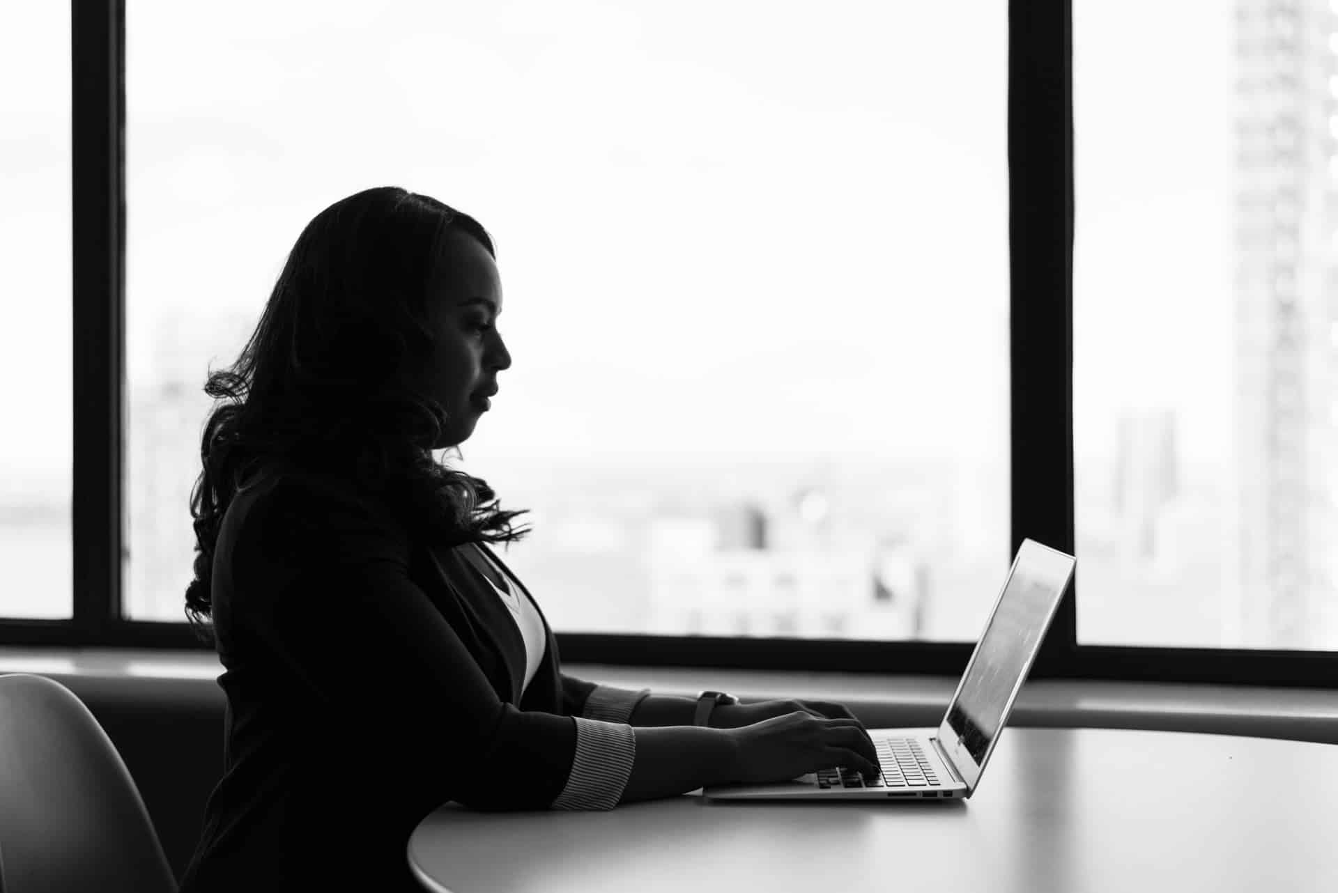 Silhouette of a Black professional woman working on a laptop at night, city skyline visible through the office window